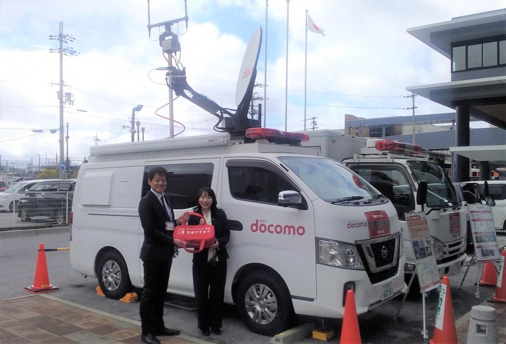 災害応援車両の前でモバイルチャージャーをもつ角田市長と三澤支店長