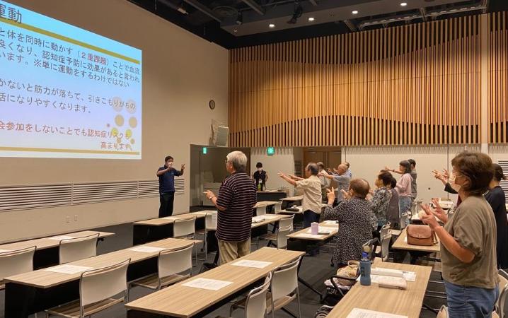 2025年に行われた認知症サポーター養成講座の写真。参加者たちが一斉に立って前にあるプロジェクタースクリーンを見ながら一斉唱和しているようす。