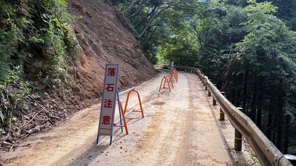 土砂撤去後のようす。土砂崩れの影響で、崖の岩肌がむき出しになっており、横の道路には通行止めしている。落石注意の看板等が設置されている。