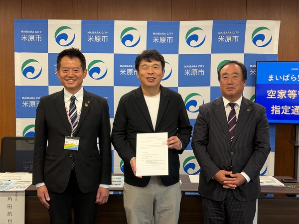 指定通知書交付式のようす。米原市ロゴマークのパネルの前に3人の男性。真ん中の男性が書類を手に持っている。