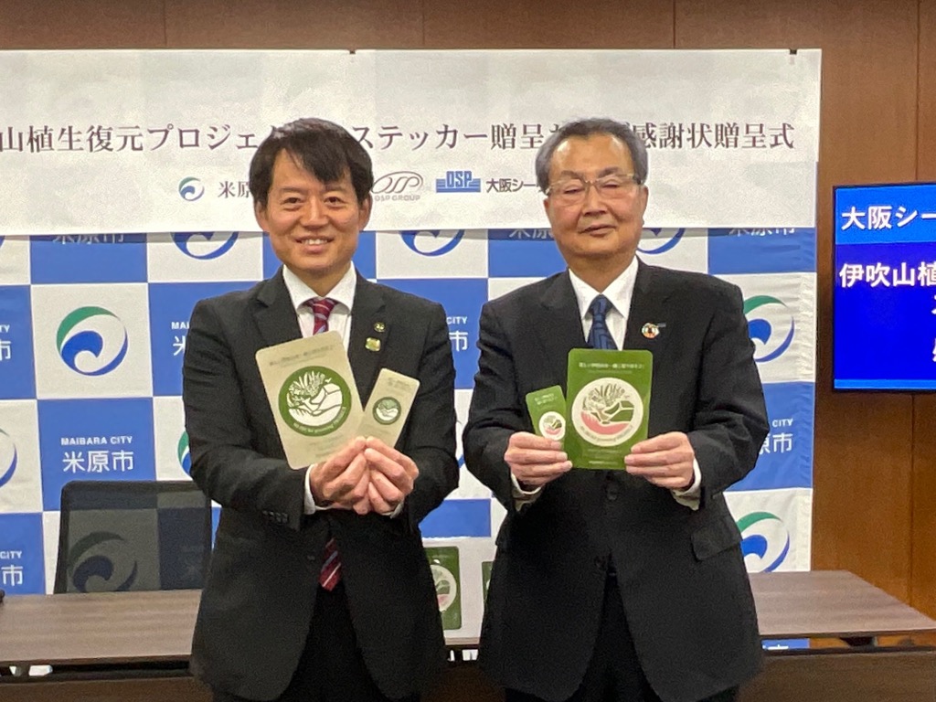 ロゴマークステッカーの寄贈写真。市長と男性の方がロゴステッカーとリーフレットを手に持っている。