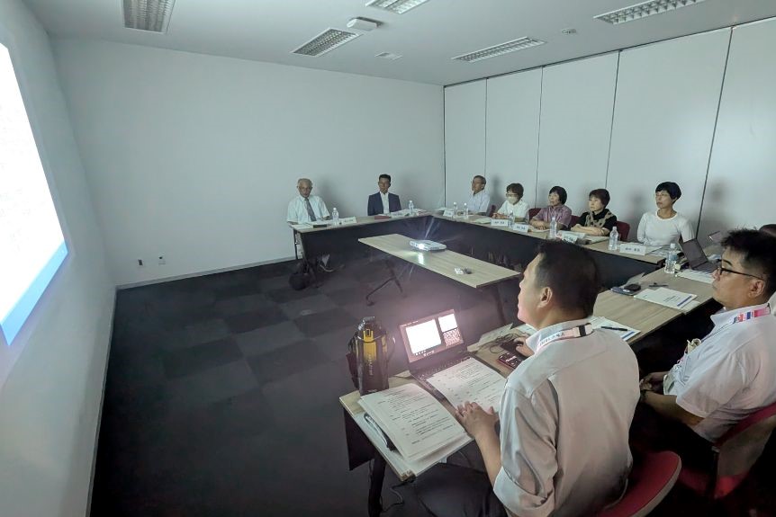 第2回審議会の開催風景。会議室の壁面に資料を投影し、事務局から委員に説明している様子。