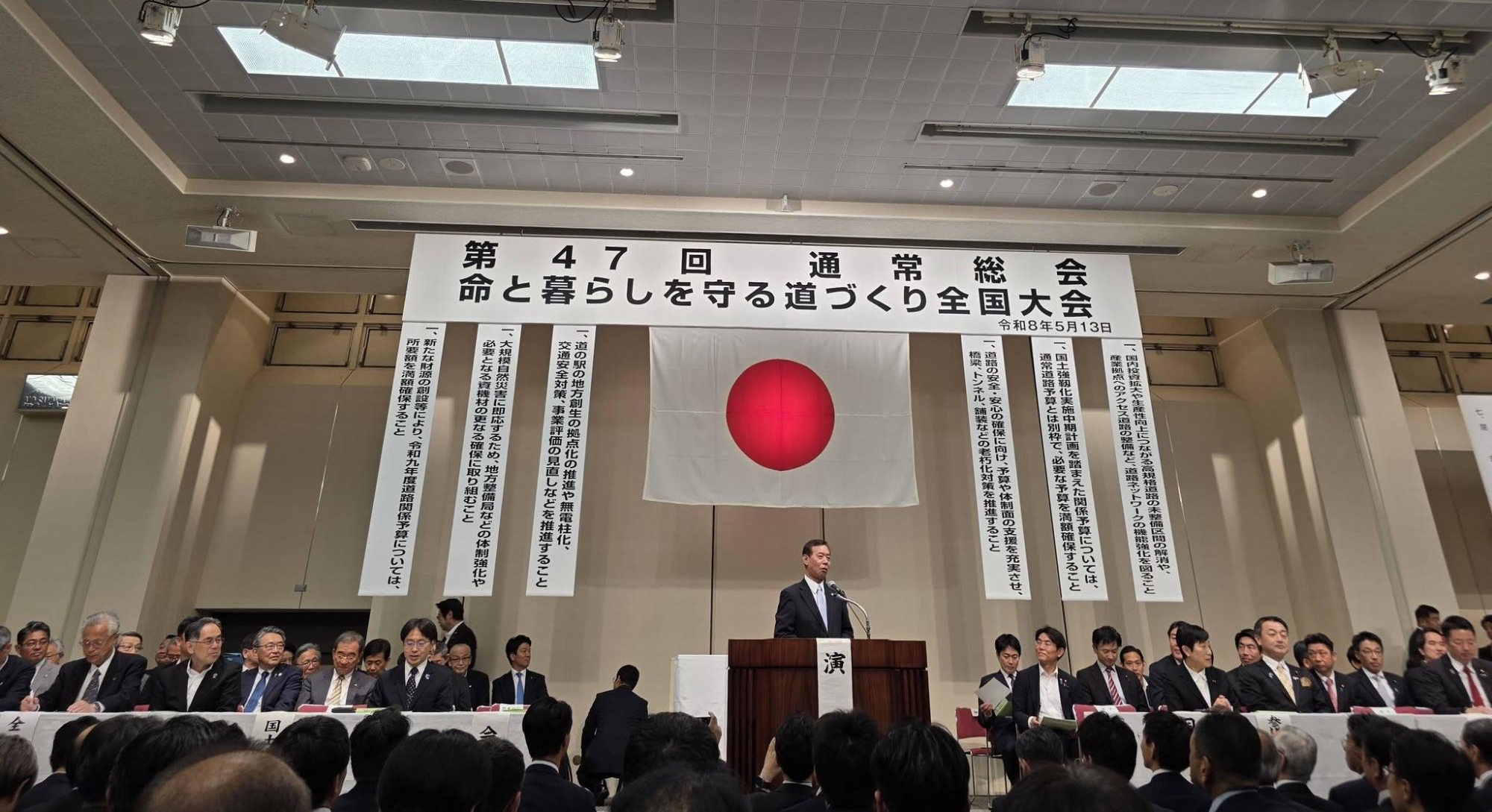 会場内で命と暮らしを守る道づくり全国大会と書かれた垂れ幕が背後にあり、壇上で挨拶されている男性の写真