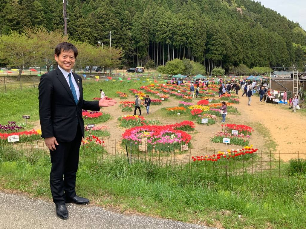 多くのチューリップが咲いており、来場者でにぎわっているようす。その少し上の場所から左手で案内しているかのようなポーズをとる市長の写真
