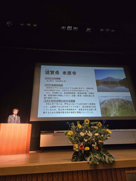 壇上にてマイクでお話されている市長の写真。後ろのプロジェクタースクリーンには、伊吹山などの写真が添えられた米原市のプロフィールが映し出されている。