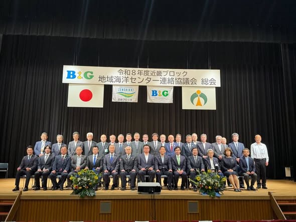 ステージ上で参加者たちが並んで立ったり座っている状態で集合写真。後ろには、令和8年度近畿ブロック地域海洋センター連絡協議会総会と書かれたパネルが吊り下げられている。