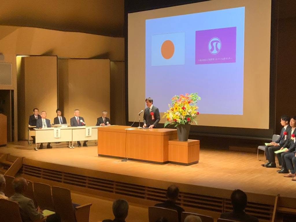 壇上にて挨拶されている市長の写真。後ろのプロジェクタースクリーンには日本、会社のシンボルマークが写しだされている。