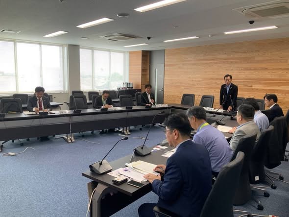 会議室にて長机が並べられており、その端っこあたりに立ってお話されている男性の写真