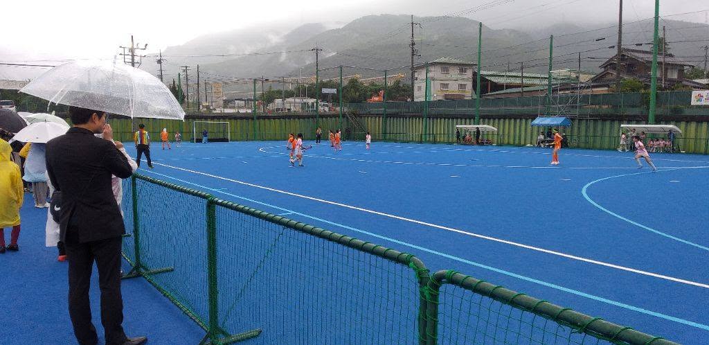 雨の中ホッケーの試合を行ってる選手たち、手前には傘をさしている観客たちの写真