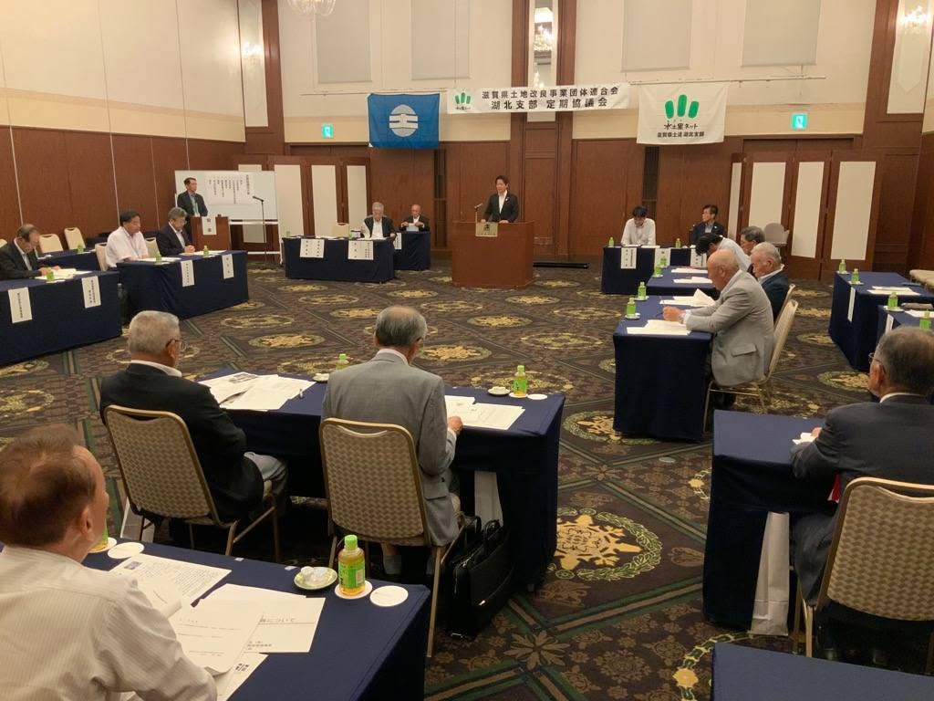 会場で、長机が並んでおりその中心の壇上でお話されている男性の写真