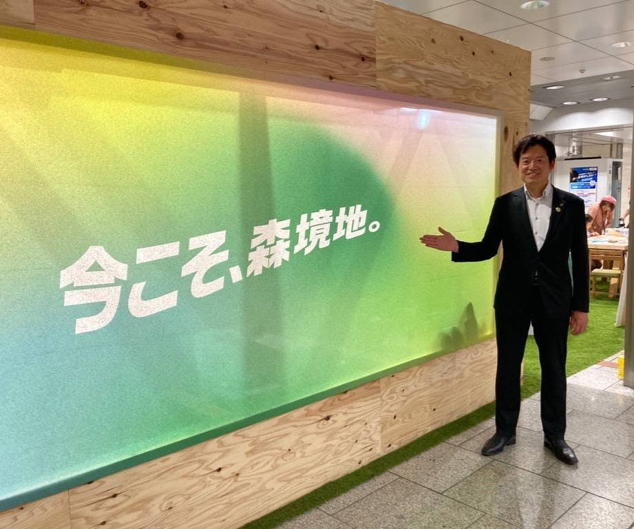 今こそ、森境地と書かれた布パネルに手のひらで指している市長の写真