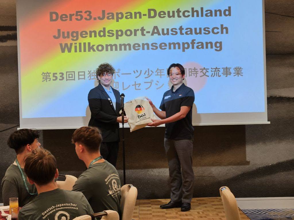 プロジェクタースクリーンが後ろにあり、その前でスーツを着た男性と市長2人でロゴマークが彩られているトートバッグを手に持っている写真
