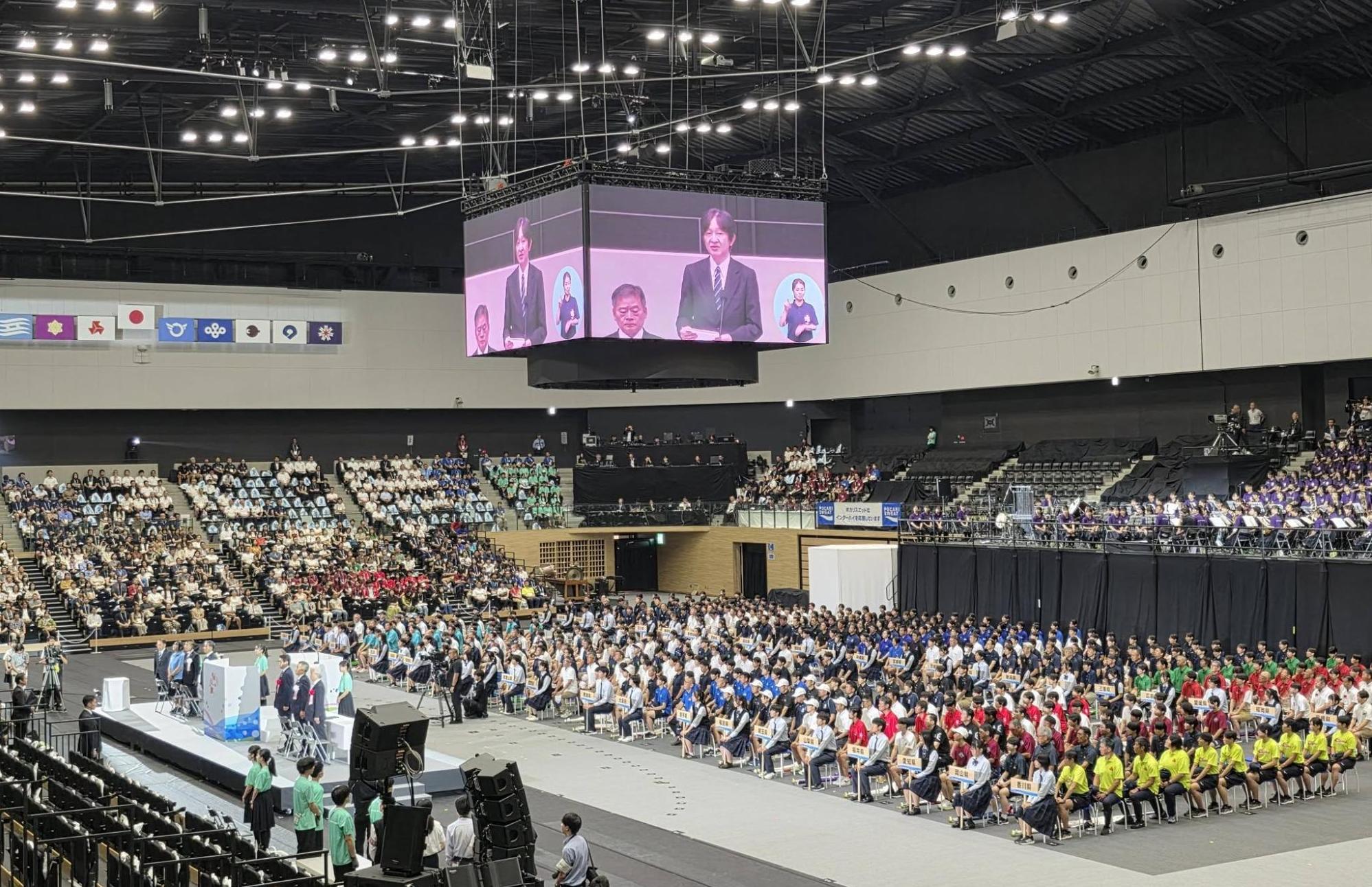 会場の中心に選手たちが固まって着席している。会場上部のテレビモニターにて挨拶されている男性が映し出されているようす。