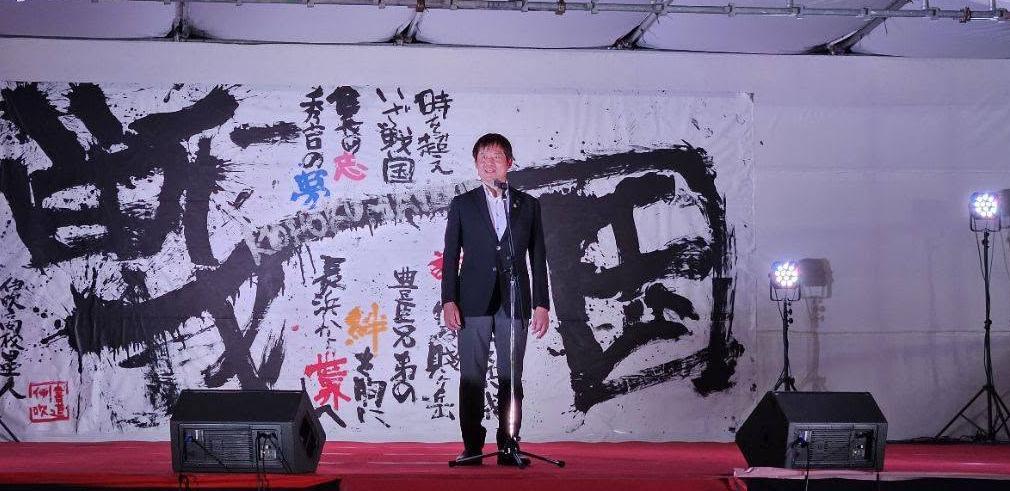 壇上にて挨拶する市長の写真。後ろには書道パフォーマンスで書かれた書が吊り下げられている。
