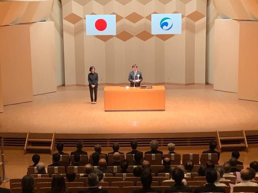 壇上にてお話されている市長の写真。後ろには国旗と市旗が吊り下げられている。