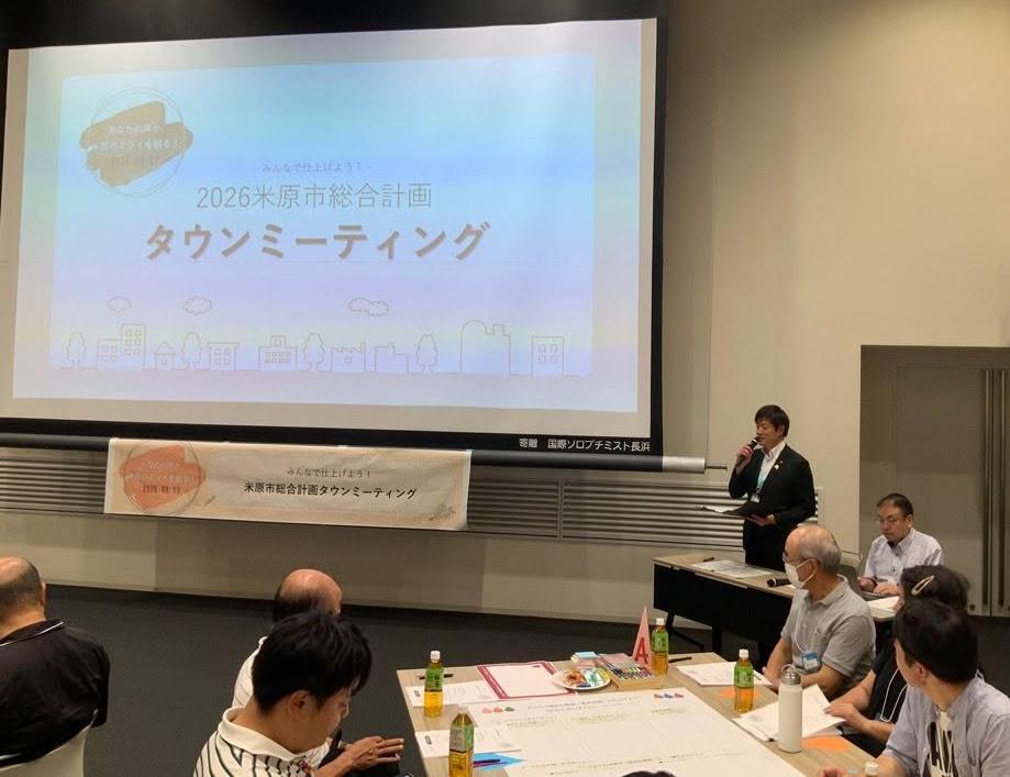 コンベンションホールにて、参加者のみなさんの前で挨拶されている市長の写真。後ろのプロジェクタースクリーンには米原市総合計画タウンミーティングと書かれている。