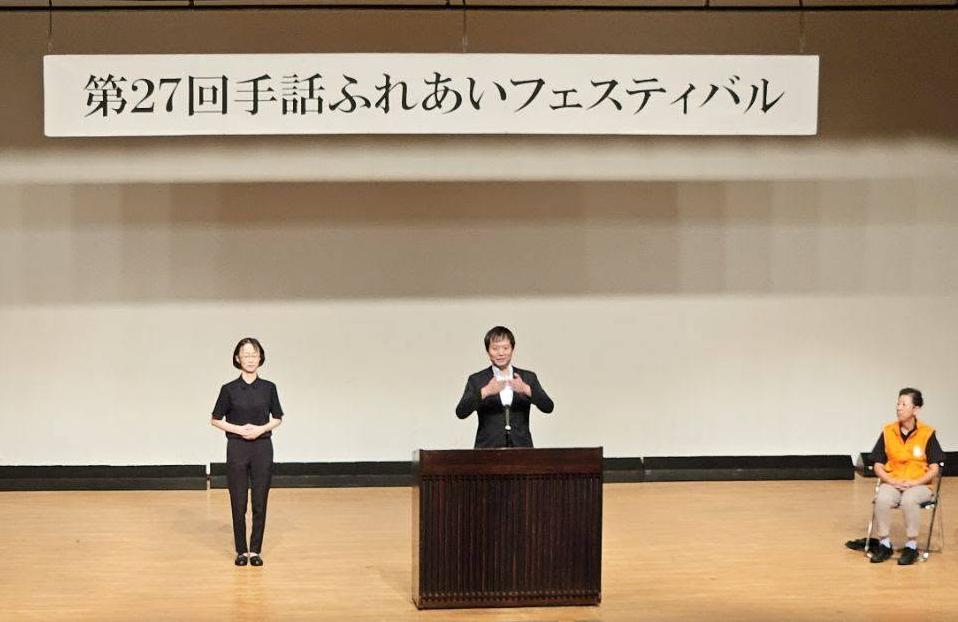 壇上にて、手話でお話されてる市長の写真。横には手話通訳者がいる。