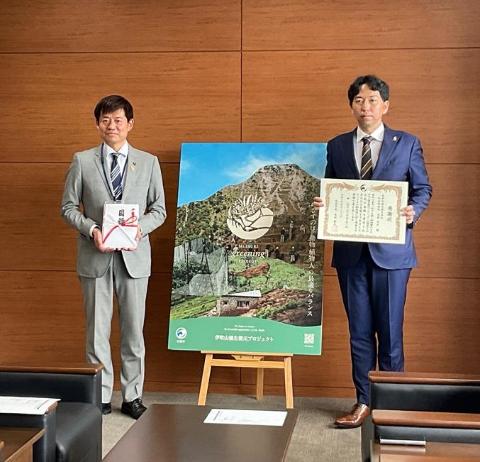 市長と奥伊吹観光会社の方の間に、奥伊吹観光会社のパネルがある。左側の市長は寄付物を手に持ち、奥伊吹観光会社の方は感謝状を手に持っている。