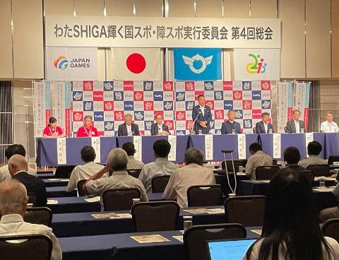 総会での様子。いくつもの長机が並んでおり、一番前には国スポ障スポ総会の横看板、パネルがありその前にマイクで話されている男性の写真