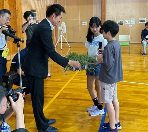 体育館で、小学生2人からイブキジャコウソウを受けとっているようす