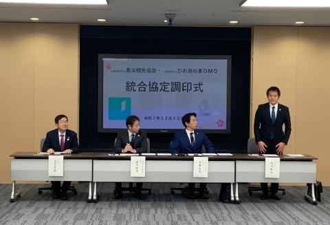 後ろに統合協定調印式と書かれたプロジェクタースクリーンがあり、その前には3人の男性が座っており、その横に立って挨拶をしている市長の写真