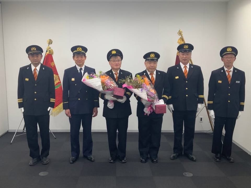 消防団退任式。市長含め6人の制服を着た男性が並んでおり、中心には花束を持っている2人がいる。