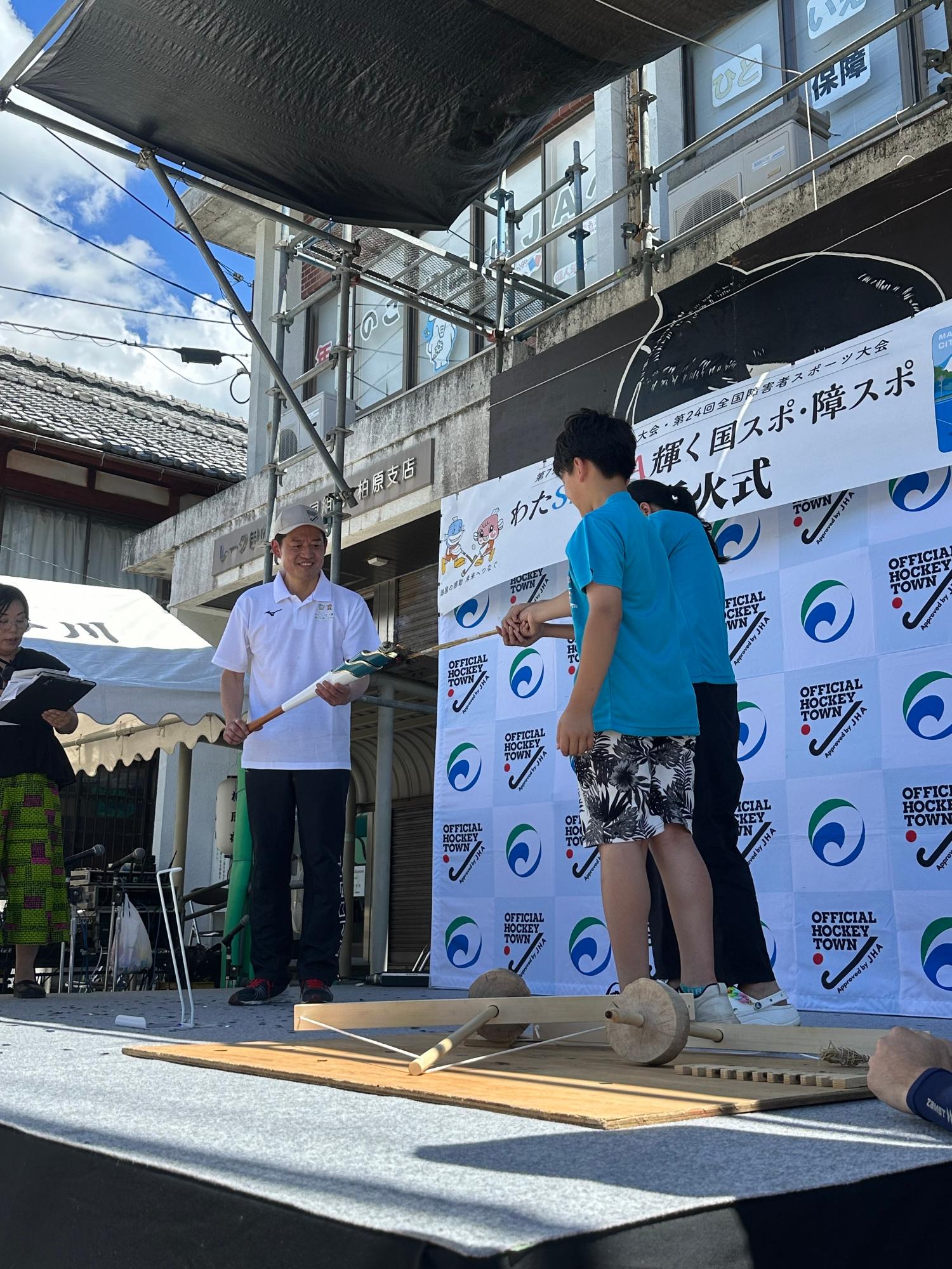 柏原やいと祭りのステージにて、採火した炬火をやいとキッズから米原市角田航也市長の炬火トーチへ移している写真。左側、米原市角田航也市長。右側、やいとキッズ2人。