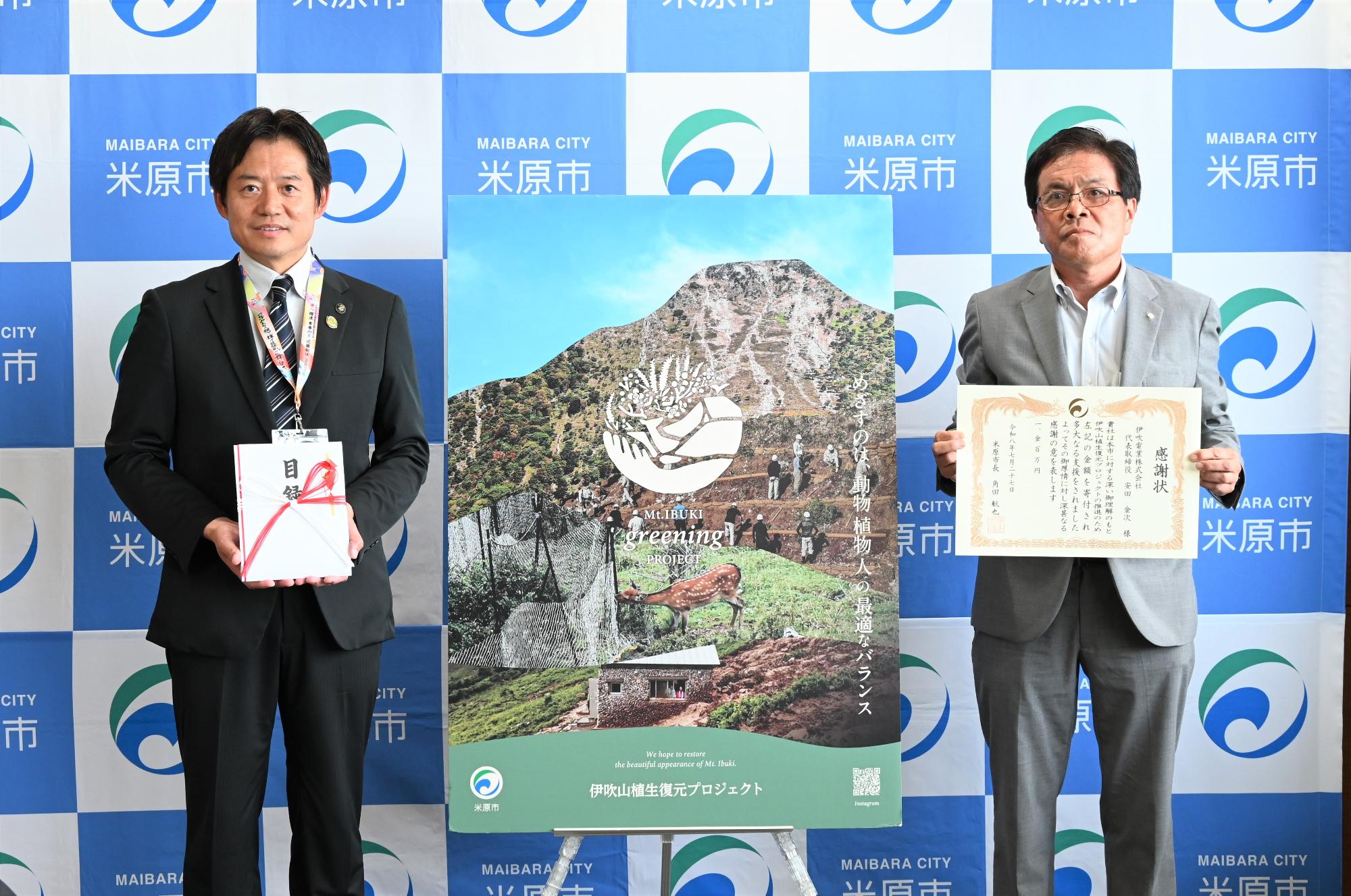 寄付贈呈式。左側に目録を手に持っている市長、その横に伊吹電業光株式会社の方が感謝状を手に持っている。2人の間にグリーニングプロジェクトのパネルが設置されている。