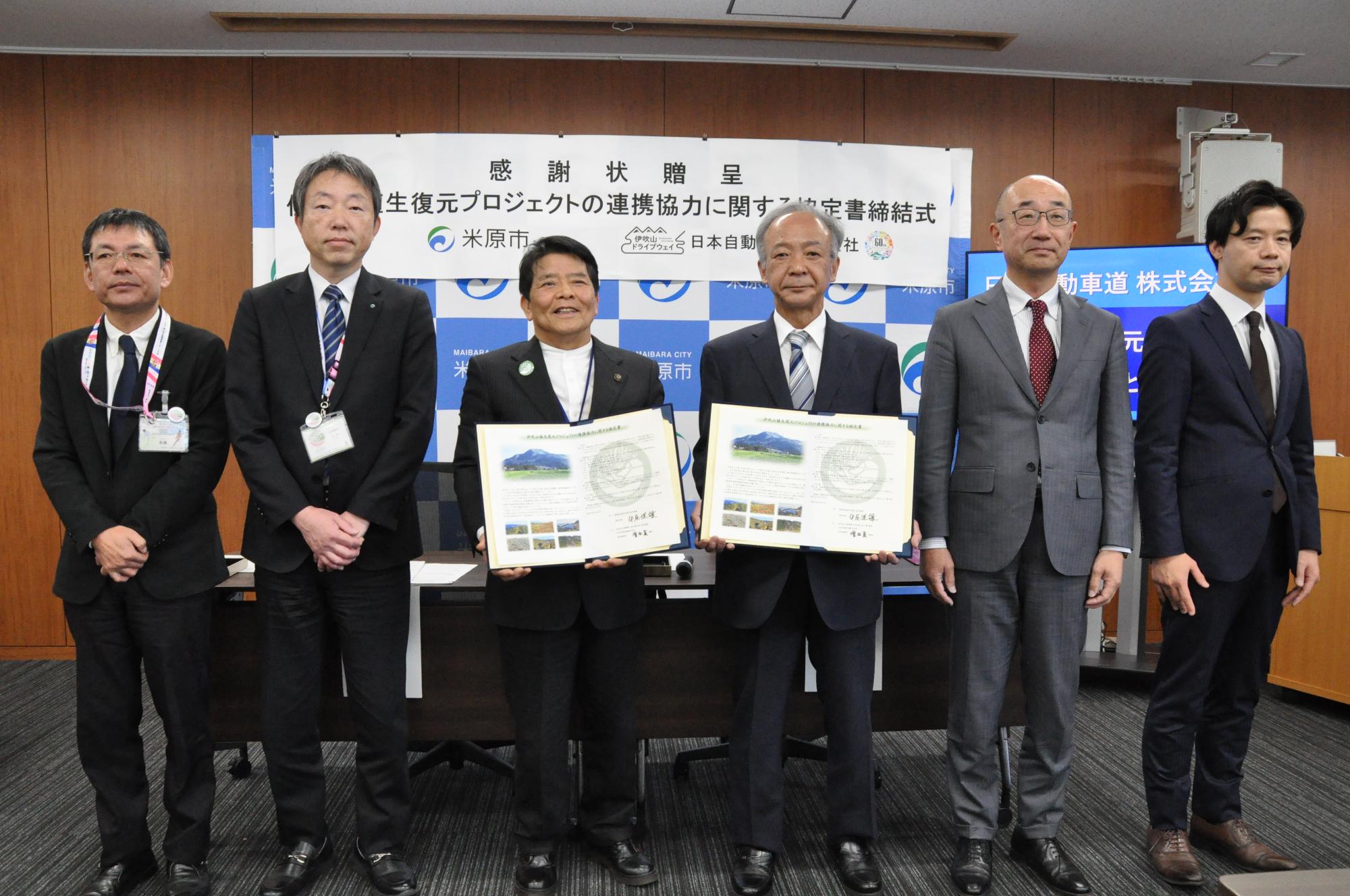 協定締結式の写真。6人の男性が並んで立っている。左側から3番目の市長、4番目の男性の方がそれぞれ協定書を手に持っている。
