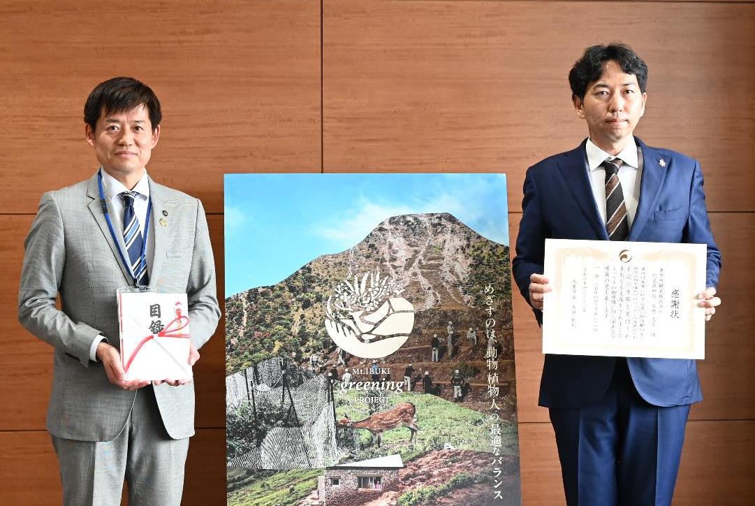 寄付贈呈式。左側に目録を手に持っている市長、その横に奥伊吹観光株式会社の方が感謝状を手に持っている。2人の間にグリーニングプロジェクトのパネルが設置されている。