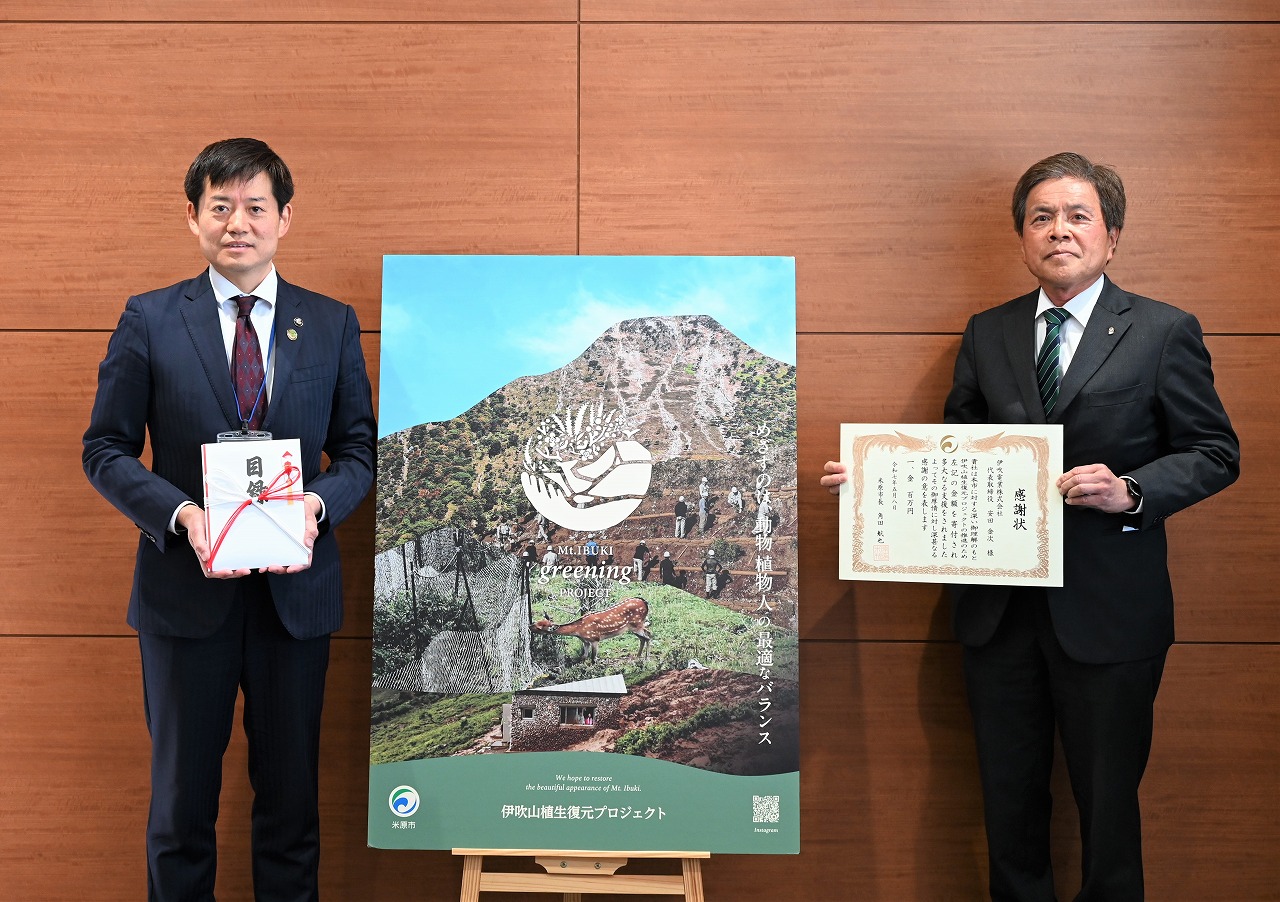 寄付贈呈式。左側に目録を手に持っている市長、その横に伊吹電業光株式会社の方が感謝状を手に持っている。2人の間にグリーニングプロジェクトのパネルが設置されている。