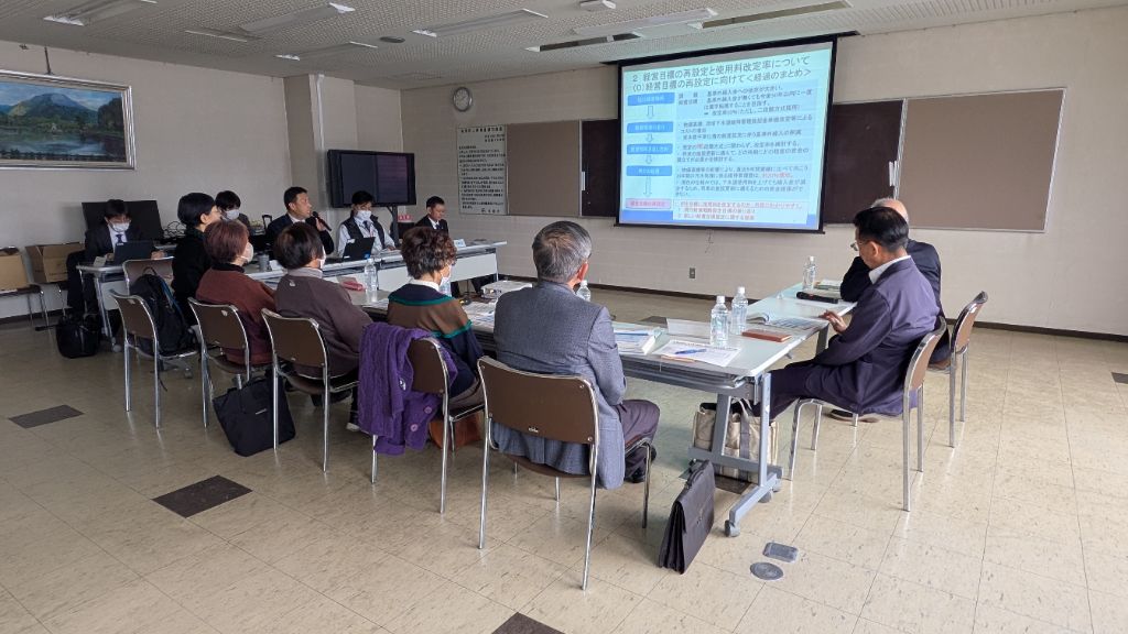 第4回審議会の開催風景。会議室正面に資料を投影し、事務局から委員に説明している様子。