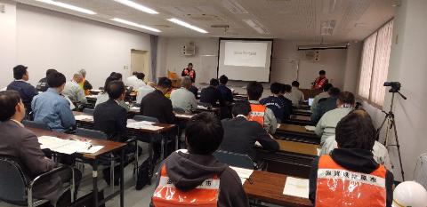 プロジェクタースクリーンに訓練内容の概要が書かれており、それを説明している職員の写真