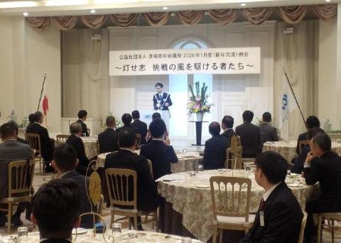 いくつかの丸机があり、大勢の人が着席しており、司会席で挨拶されている男性の写真。