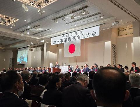 ホール内にて、ステージ上は治水事業促進全国大会のスローガンが掲げられており、大勢の人が出席している写真