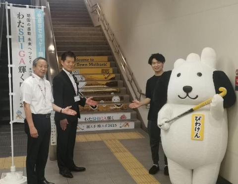 階段装飾の前に、市職員、市長、ベストアンサー株式会社の方、マスコットキャラクターびわけんの着ぐるみと並んで撮影
