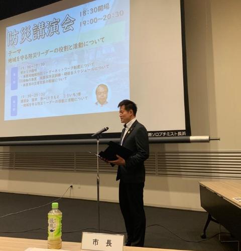 後ろにプロジェクタースクリーンがあり、スタンドマイクで挨拶をされている市長の写真