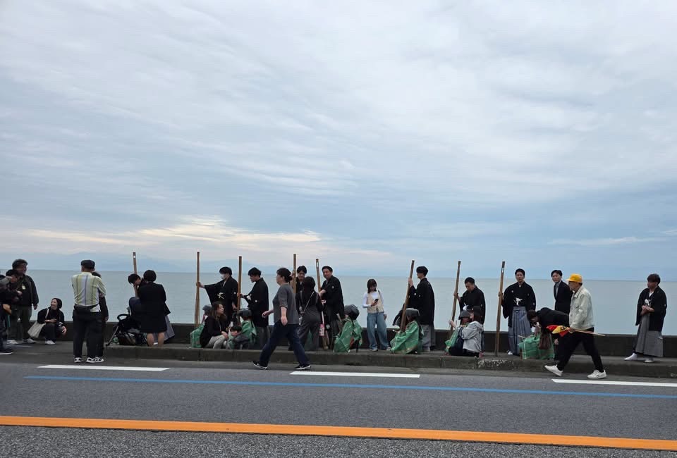 鍋冠祭のようす。鍋のような形をしている被り物を被っているこどもと黒紋付を着ている男性の集団が練り歩いているようすの写真