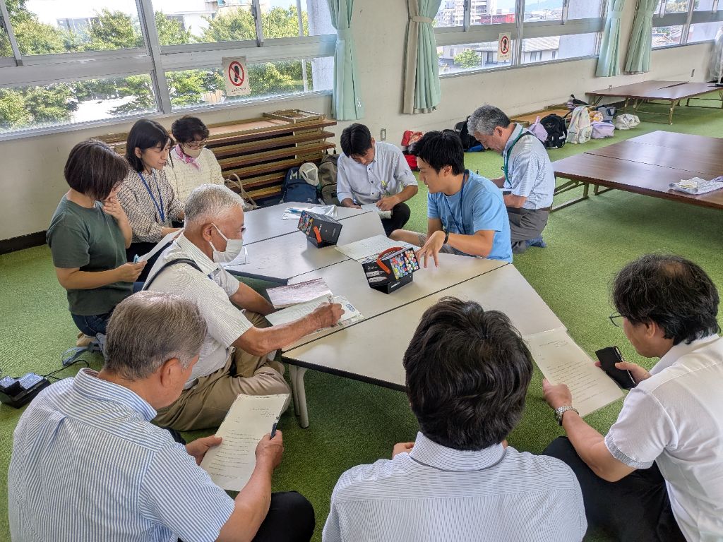 学校の一室で講師が事業の説明をし、周囲で社会教育委員が記録をしている様子。