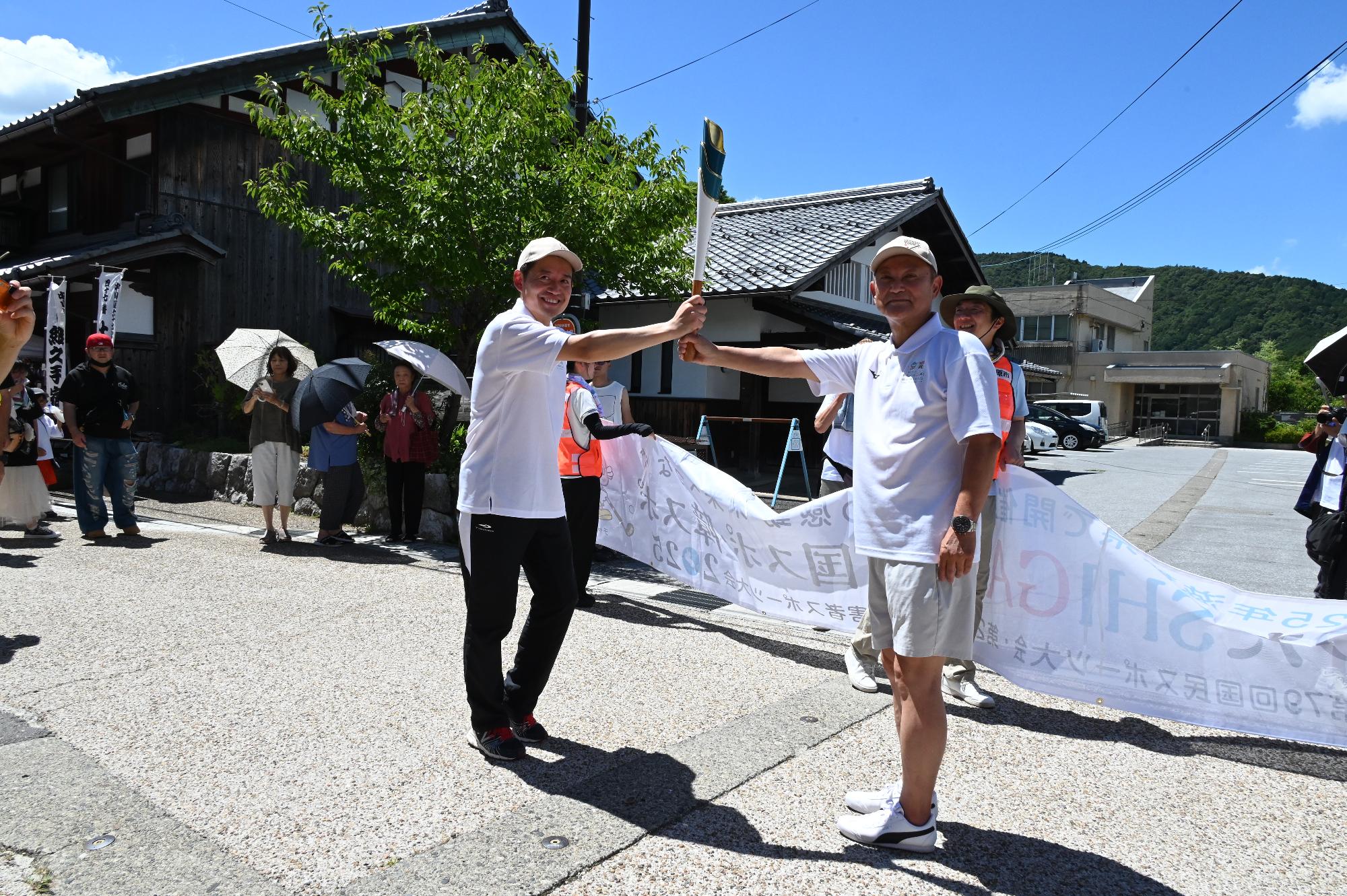 炬火リレーでトーチを受け渡す写真。左側、米原市の角田航也市長。右側、米原市議会議員の山本克己議員。