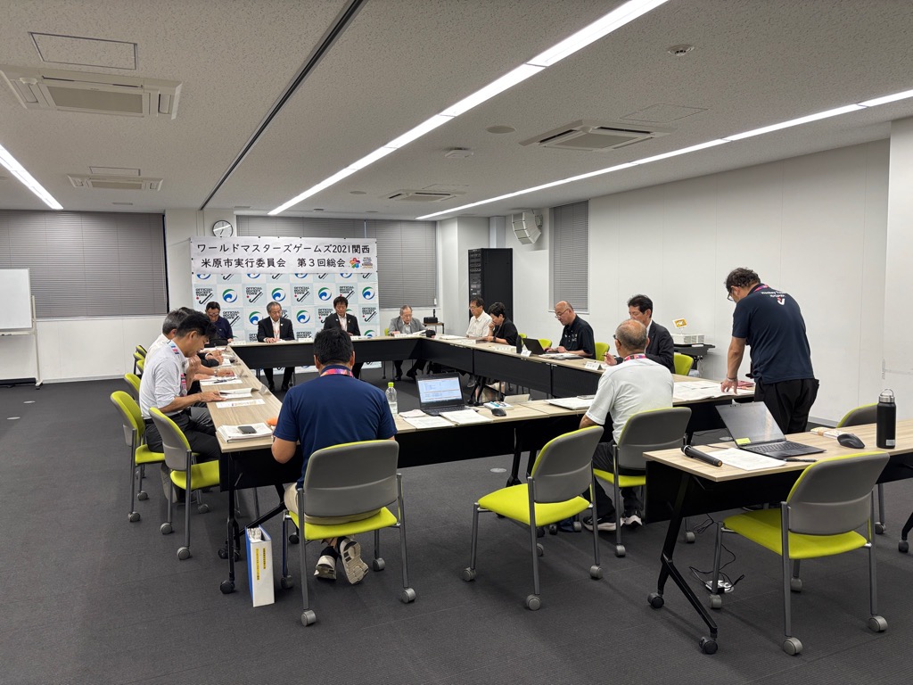 四角に囲んだ机があり職員が着席している。手前の席に立ってお話している男性の写真